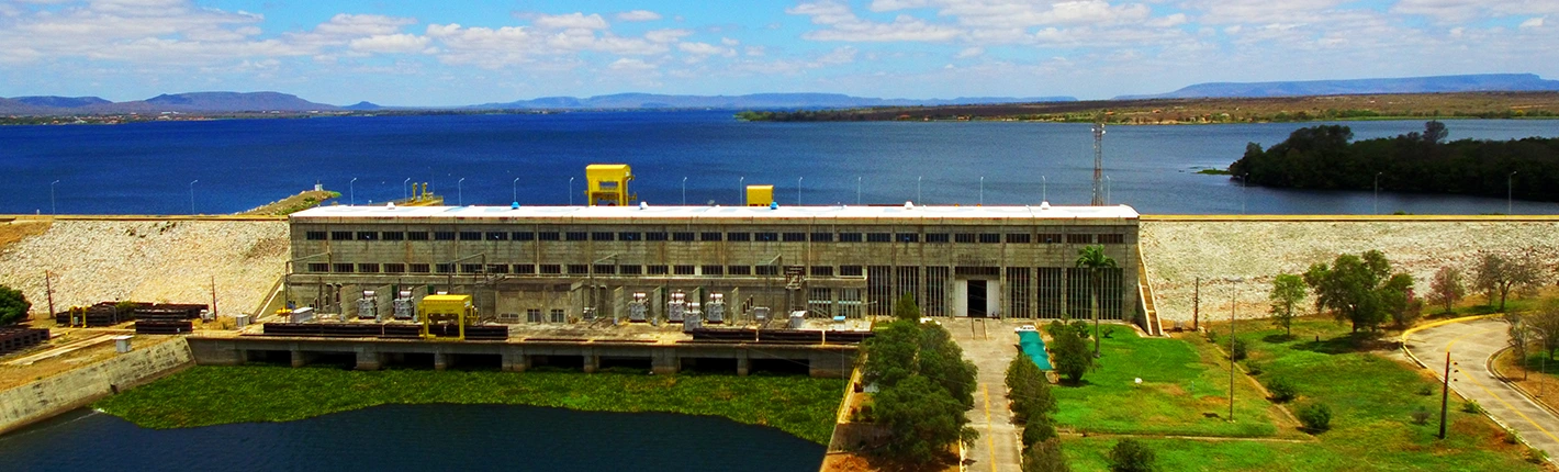 Usina hidrelétrica à beira de grande reservatório, com prédio operacional, áreas verdes e céu azul com nuvens ao fundo.