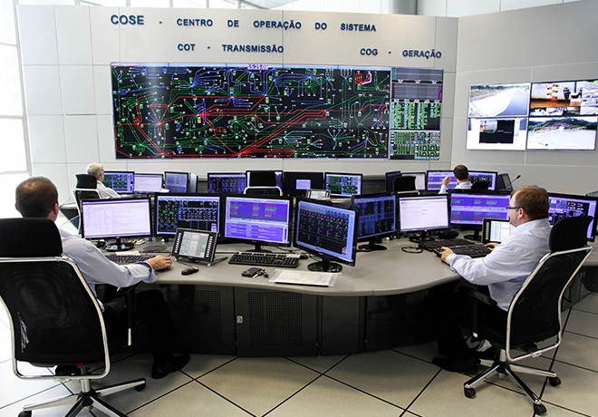 Sala de controle de sistema elétrico com operadores em computadores e vários monitores, incluindo um grande painel com diagramas de rede e dados de transmissão.