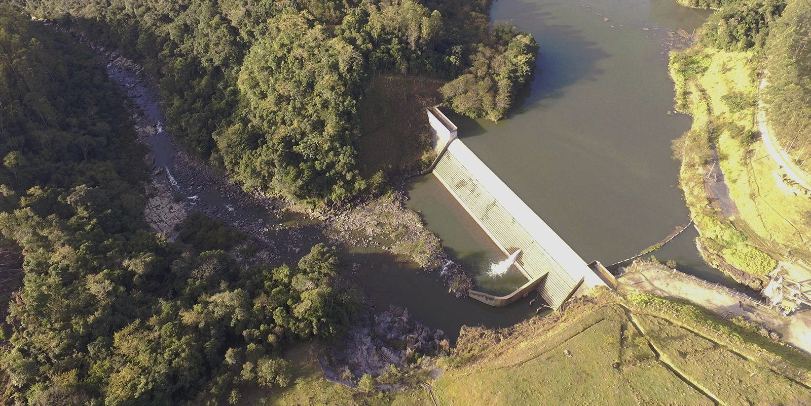 Vista aérea de uma pequena barragem hidrelétrica com vertedouro em degraus, liberando água em fluxo contínuo para um rio estreito que serpenteia entre áreas de vegetação densa, cercada por relevo irregular, margem rochosa e uma área aberta com gramado e acesso lateral.