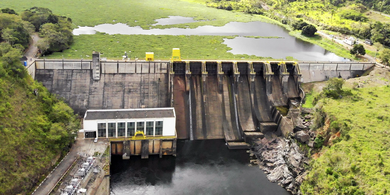 Vista frontal de uma usina hidrelétrica com barragem de concreto e vertedouro equipado com múltiplas comportas, casa de força ao lado e água escoando de forma controlada, cercada por vegetação densa e pequenas áreas alagadas ao fundo.