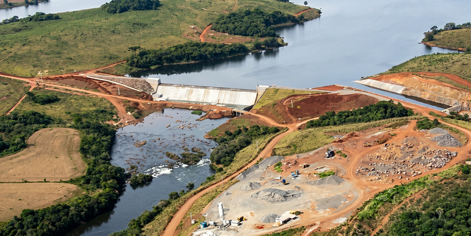 Vista aérea de uma pequena barragem hidrelétrica com vertedouro central, separando um reservatório azul profundo com ilhas vegetadas de um canal a jusante, cercada por áreas de solo avermelhado, estradas de terra e vegetação dispersa em relevo suave.