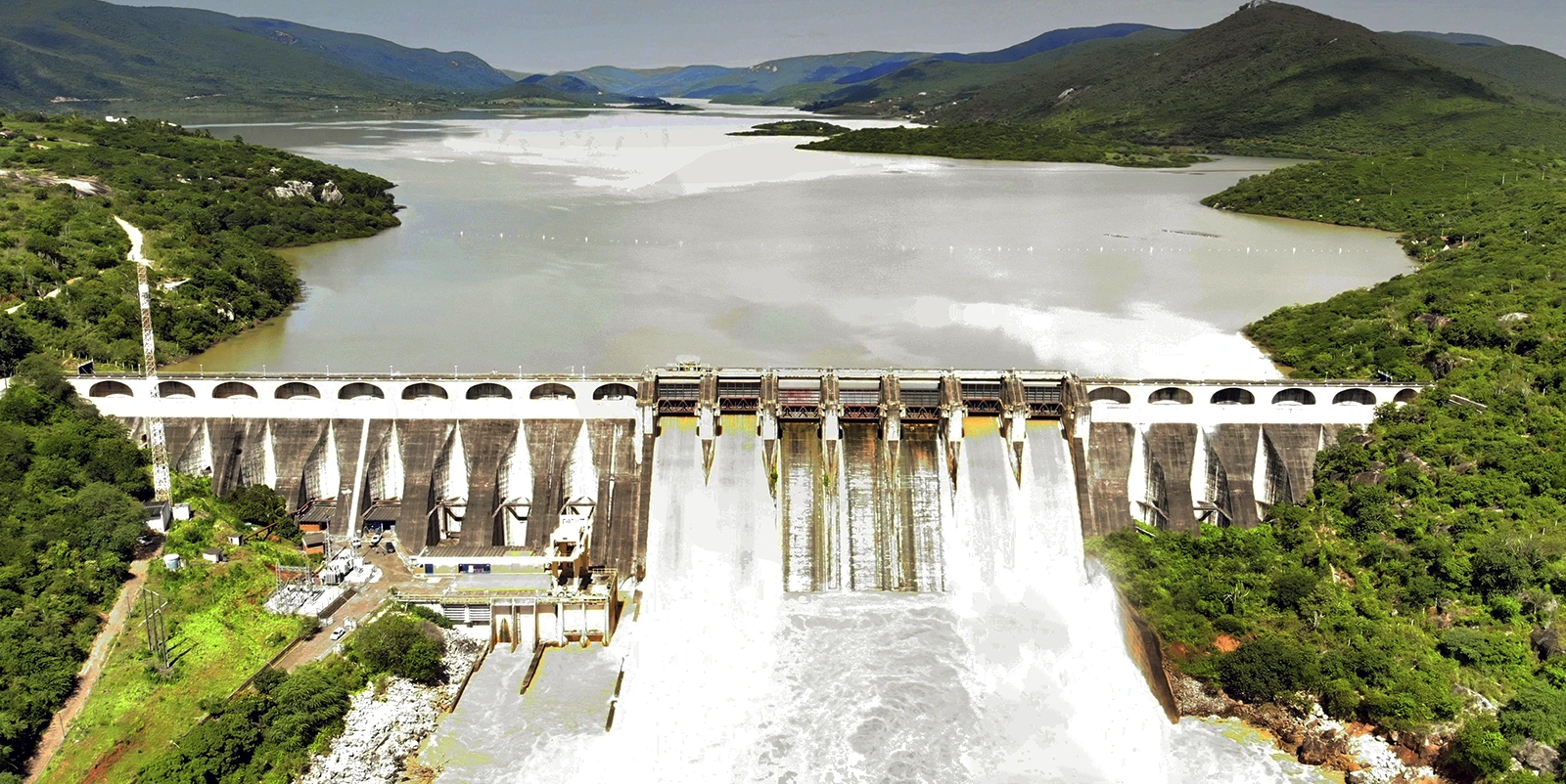 Vista frontal de uma barragem hidrelétrica extensa, com vertedouro em operação liberando grande volume de água em cascata, reservatório amplo ao fundo cercado por morros verdes e vegetação densa, sob céu claro.