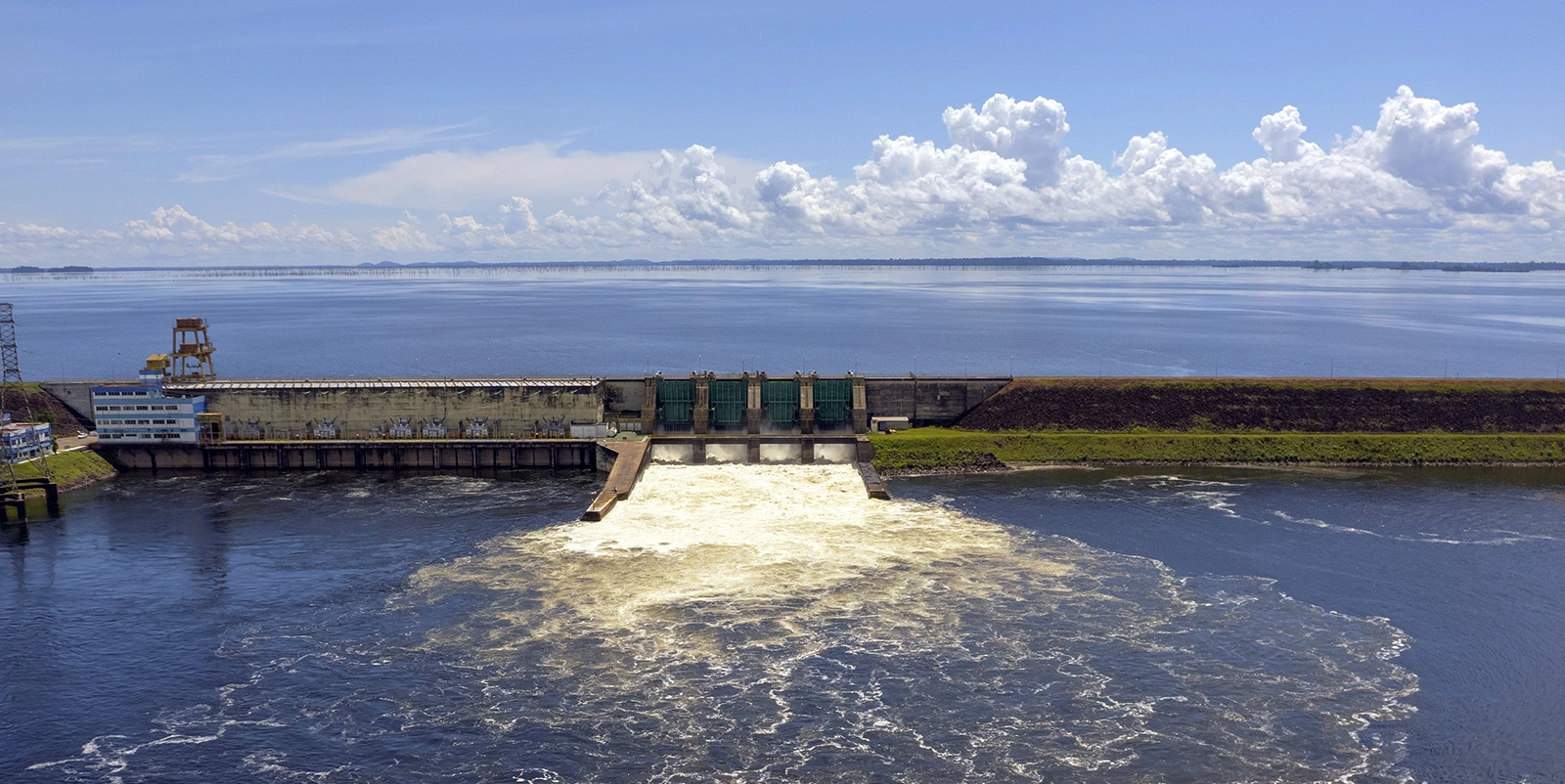 Vista frontal de uma barragem hidrelétrica com três comportas centrais liberando água em fluxo contínuo que se espalha pelo rio a jusante, com casa de força e torre de operação à esquerda, reservatório amplo e calmo ao fundo e céu claro com nuvens dispersas.