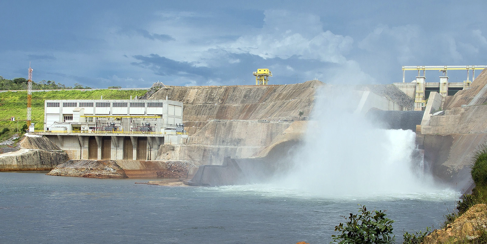 Usina hidrelétrica em operação com vertedouro liberando grande volume de água em cascata, formando névoa sobre o rio, ao lado de estruturas industriais e paredão de terra sob céu parcialmente nublado.