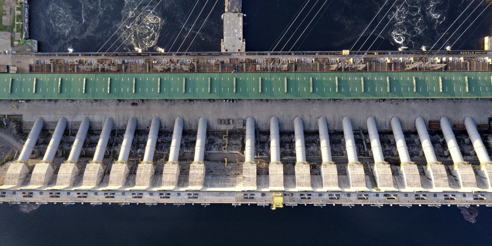 Vista aérea em perspectiva vertical de uma usina hidrelétrica com longa casa de força de cobertura verde ao centro, alinhada a uma série de grandes condutos metálicos inclinados dispostos em sequência, conectando a barragem ao canal inferior, com água escura em ambos os lados, estruturas industriais, passarelas e equipamentos distribuídos ao longo da instalação.