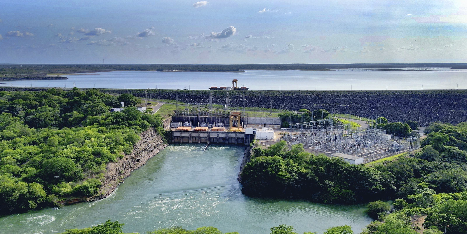 Vista frontal de um complexo hidrelétrico com barragem baixa e comportas sobre um canal estreito, acompanhado de subestação elétrica com torres e equipamentos, cercado por vegetação densa nas encostas e um grande reservatório ao fundo protegido por dique elevado.