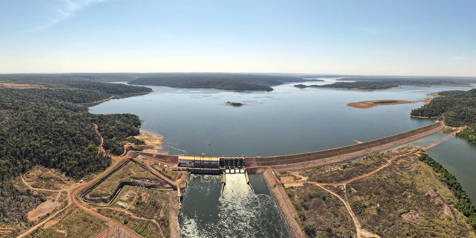 Vista aérea ampla de um sistema hidrelétrico com barragem longa formando um reservatório extenso que se ramifica entre áreas de vegetação densa e ilhas, com vertedouro ao centro liberando água para o canal a jusante, cercado por estradas de terra, áreas abertas e relevo suave sob céu claro.