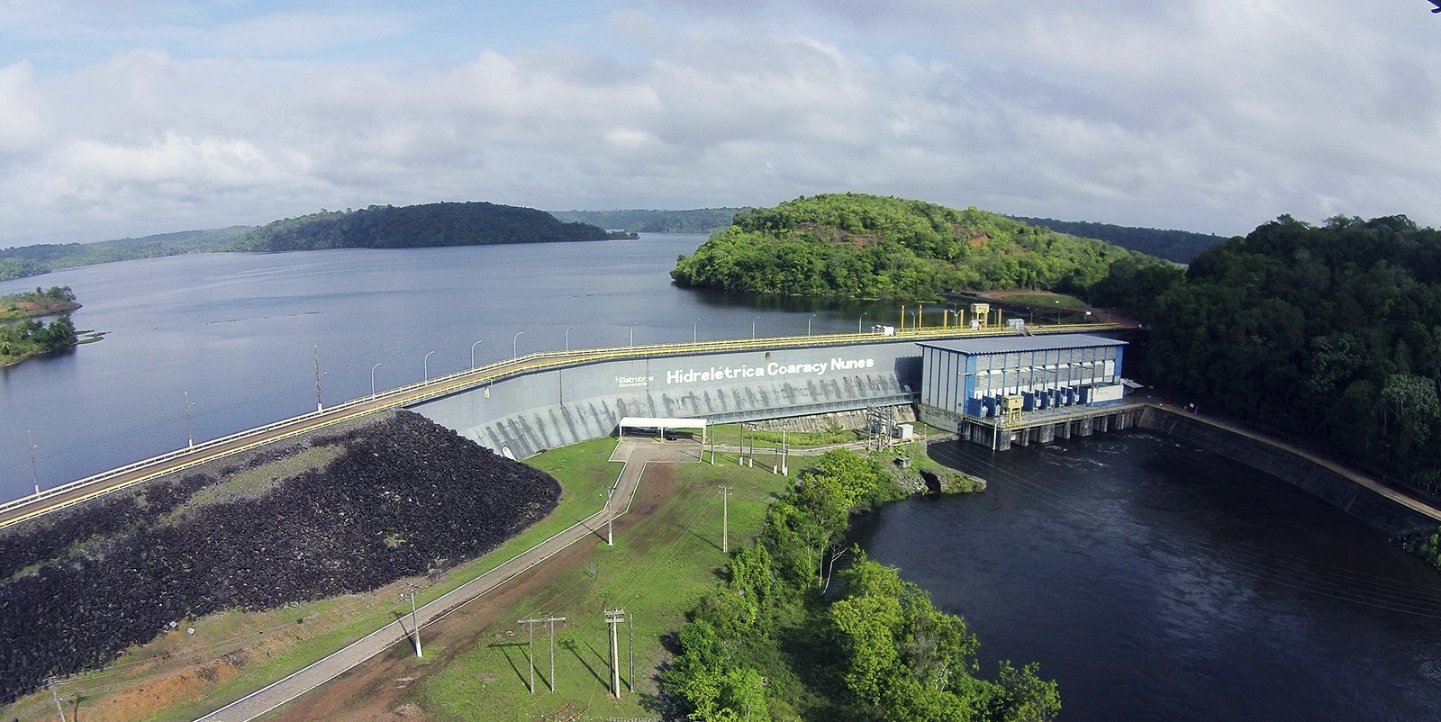 Vista lateral de uma barragem hidrelétrica de enrocamento com casa de força adjacente, onde se lê “Hidrelétrica Coaracy Nunes” na estrutura, cercada por um amplo reservatório e áreas de vegetação densa, com estrada sobre o coroamento sob céu parcialmente nublado.