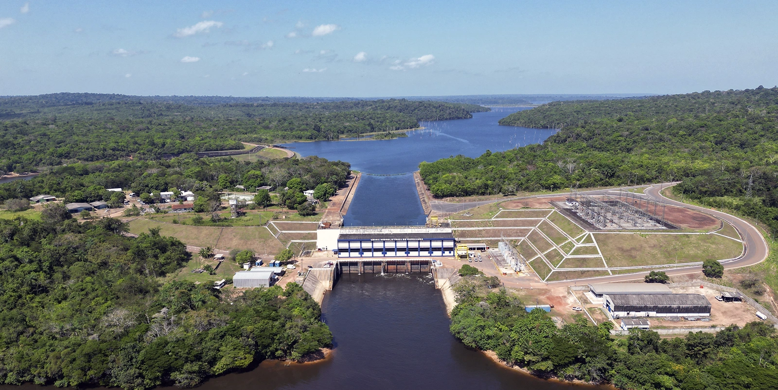Vista frontal de uma usina hidrelétrica com casa de força retangular sobre o canal principal, conectando um reservatório amplo ao fundo a um rio mais estreito a jusante, cercada por vegetação densa, estradas de acesso e uma subestação elétrica adjacente à direita.