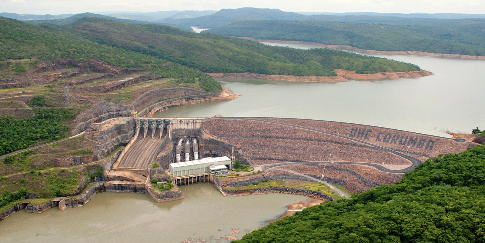 Vista aérea de uma usina hidrelétrica integrada a reservatório amplo, cercada por áreas verdes e montanhosas, com barragem de enrocamento destacando o nome “UHE Corumbá”.