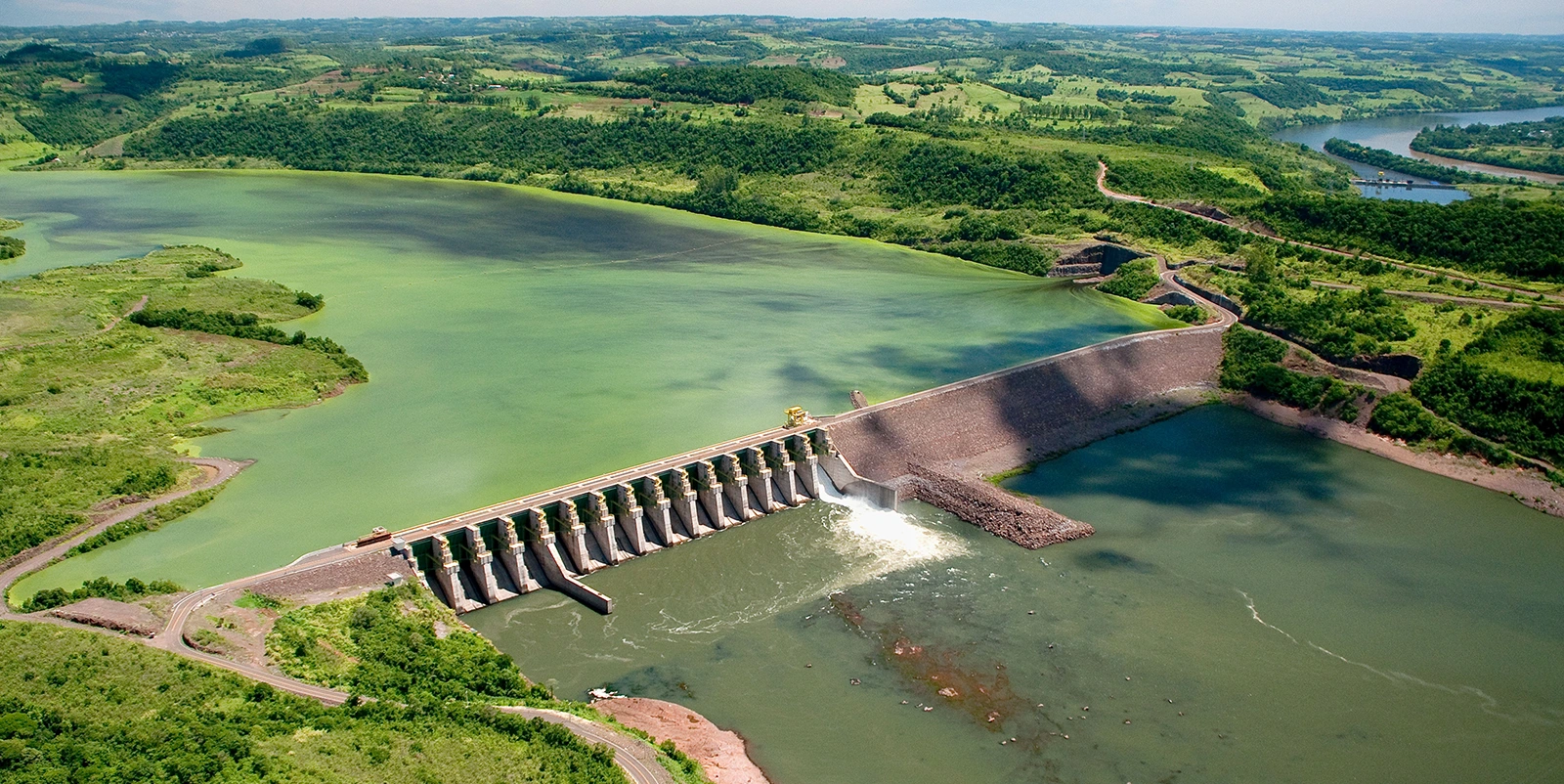 Barragem hidrelétrica em área rural vista de cima, com série de comportas liberando água, reservatório extenso e paisagem verde ao redor com colinas e estradas.