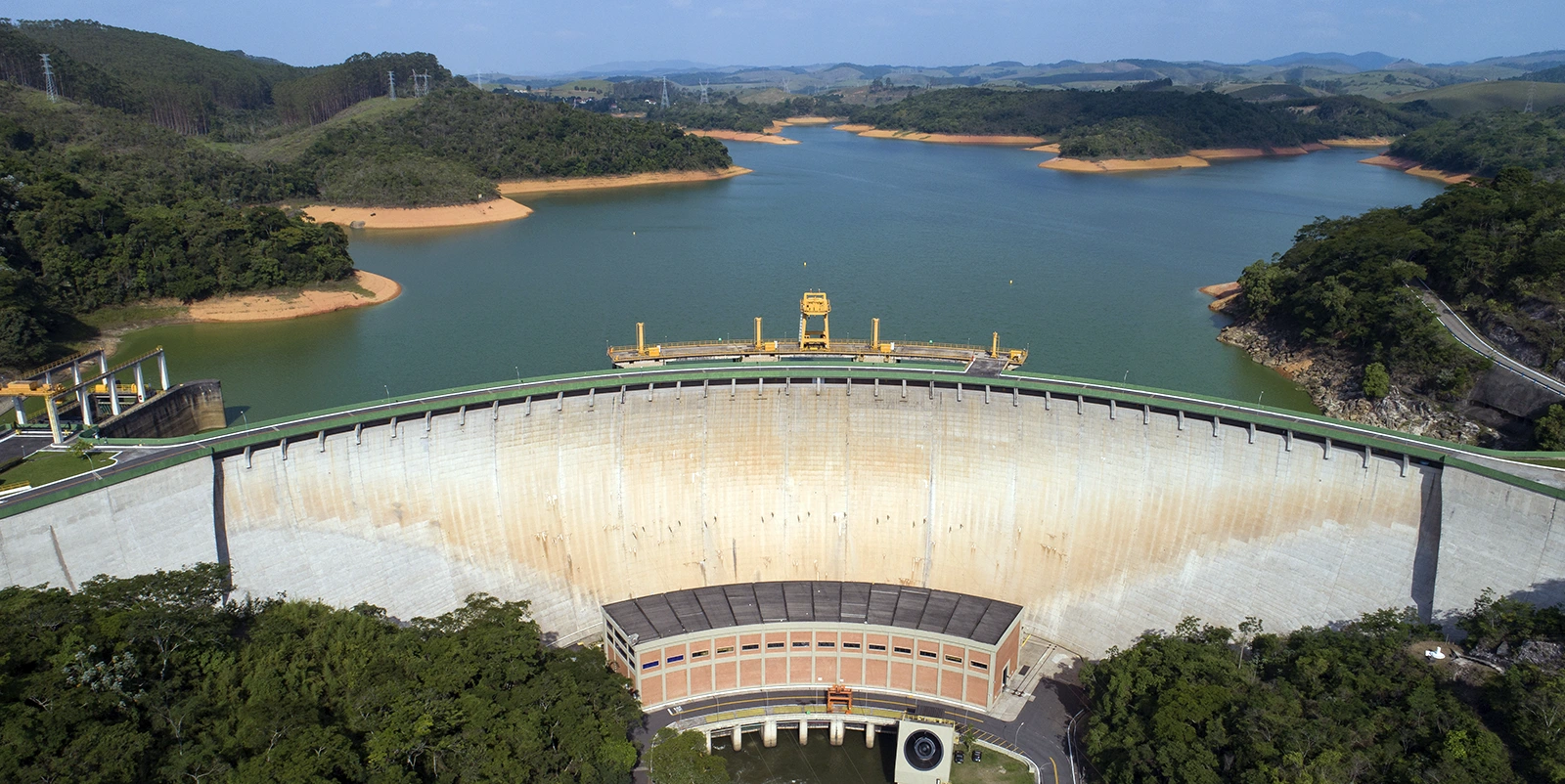 Vista frontal de uma barragem hidrelétrica em arco com grande reservatório ao fundo, cercada por vegetação densa e colinas verdes sob céu claro.