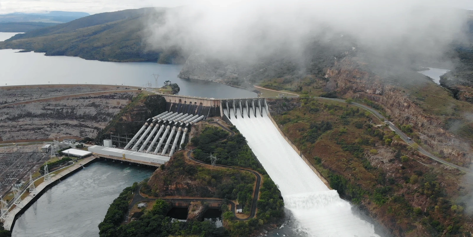 Vista aérea de um complexo hidrelétrico em região montanhosa sob neblina, com barragem e vertedouro amplo liberando forte volume de água, além de múltiplos condutos inclinados conectados à casa de força, cercados por vegetação densa, estradas sinuosas e reservatório ao fundo.