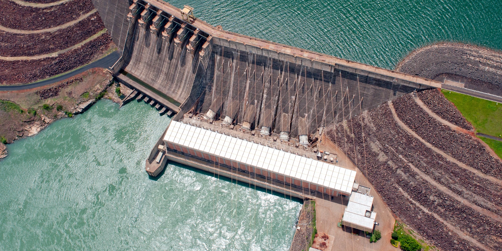 Vista aérea de uma barragem hidrelétrica de concreto com vertedouro em declive e múltiplos canais de dissipação, conectada a uma casa de força alongada na base, onde a água escoa para um canal amplo de cor esverdeada, cercada por taludes de enrocamento, áreas de terra exposta e reservatório ao topo.