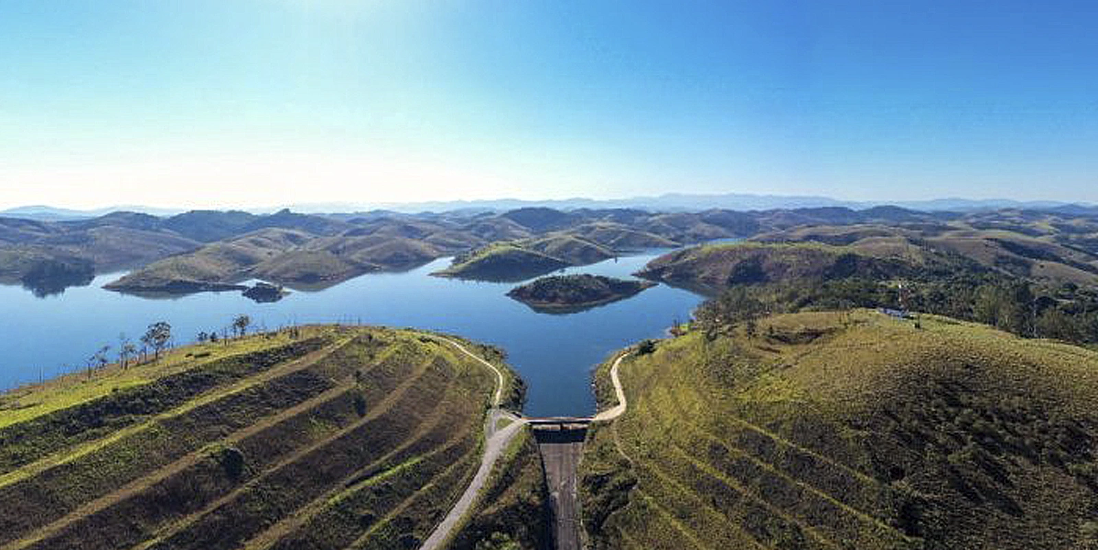 Vista aérea de um grande reservatório hidrelétrico cercado por morros e vegetação, com pequenas ilhas e estradas sinuosas nas encostas, sob céu limpo e ampla paisagem natural.