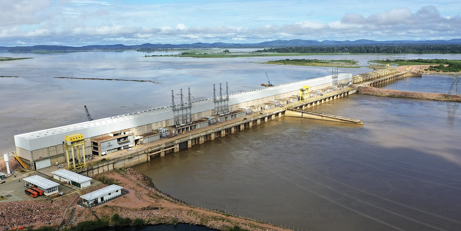 Vista aérea de uma usina hidrelétrica de grande porte com casa de força alongada sobre a barragem, equipada com guindastes amarelos e estruturas técnicas ao longo da cobertura, conectada a um extenso reservatório e a um canal de jusante, cercada por áreas de vegetação, margens rochosas e obras de apoio sob céu parcialmente nublado.