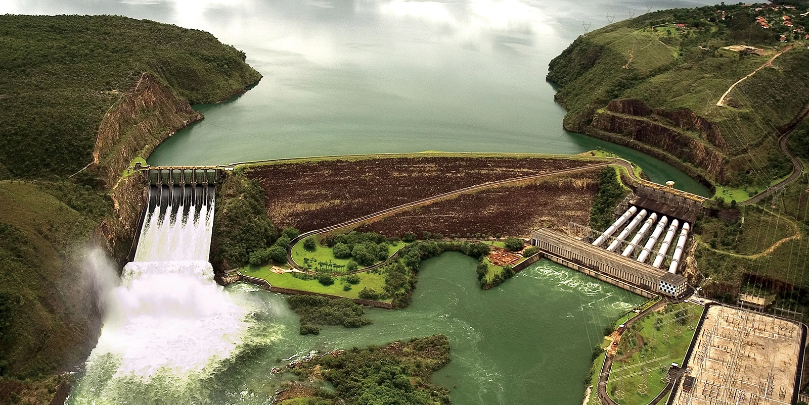 Vista aérea de um complexo hidrelétrico com barragem de enrocamento ao centro separando um amplo reservatório e o rio a jusante, vertedouro à esquerda liberando grande volume de água em queda contínua, e casa de força à direita conectada por vários condutos inclinados, além de subestação elétrica e linhas de transmissão, tudo cercado por colinas verdes e margens recortadas.