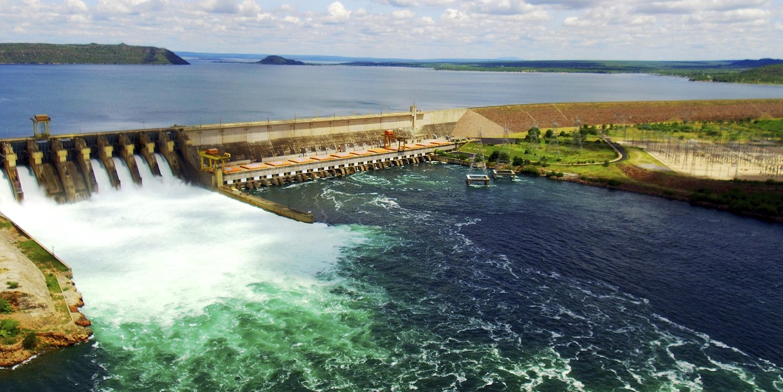 Vista ampla de uma barragem hidrelétrica com múltiplas comportas abertas, liberando grande volume de água, ao lado de uma casa de força linear, com amplo reservatório ao fundo e paisagem de morros e céu com nuvens dispersas.