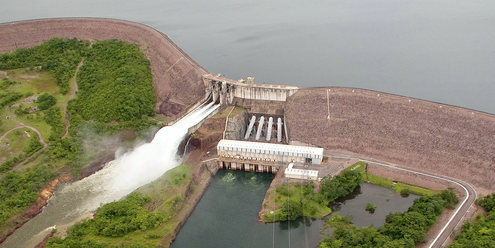Vista aérea de uma usina hidrelétrica com barragem de enrocamento e casa de força central, onde o vertedouro libera grande volume de água, cercada por vegetação densa, reservatório amplo e linhas de transmissão ao redor.
