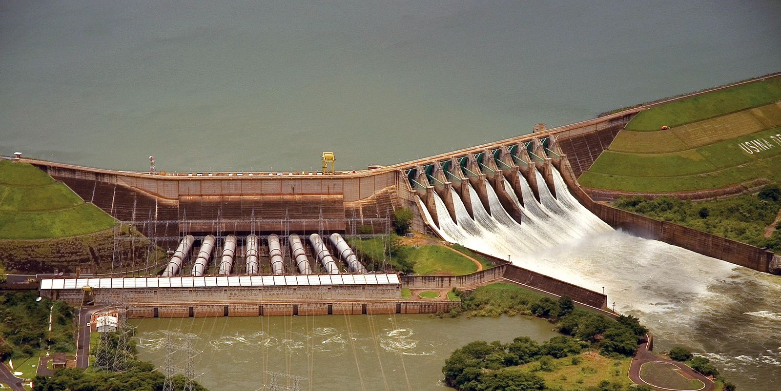 Vista aérea de uma barragem hidrelétrica com vertedouro em operação e reservatório amplo ao fundo, cercada por áreas verdes