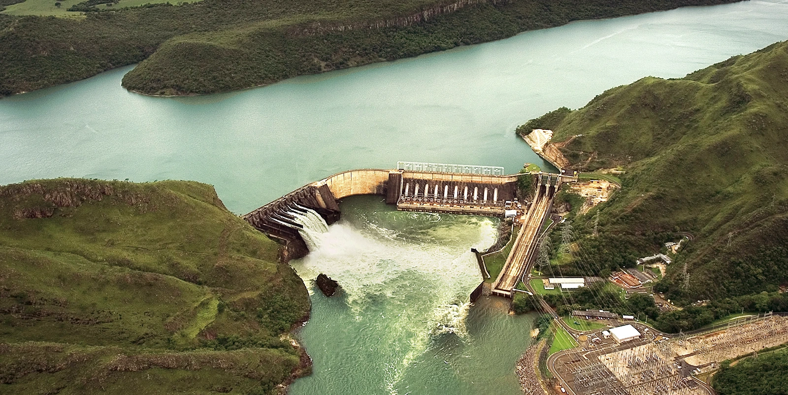 Vista aérea de uma barragem hidrelétrica instalada em um vale cercado por colinas verdes, com vertedouro alcançando o rio a jusante em forte fluxo de água, casa de força adjacente e subestação elétrica com linhas de transmissão visíveis em primeiro plano, além de um extenso reservatório ao fundo.