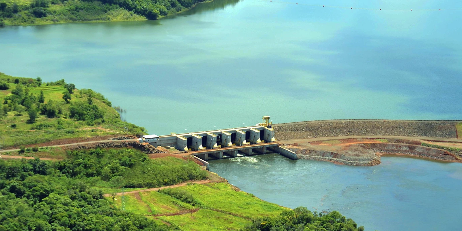 Barragem hidrelétrica vista de cima, com várias comportas ao centro, grande reservatório ao fundo e vegetação densa ao redor, incluindo áreas de mata e campos abertos.
