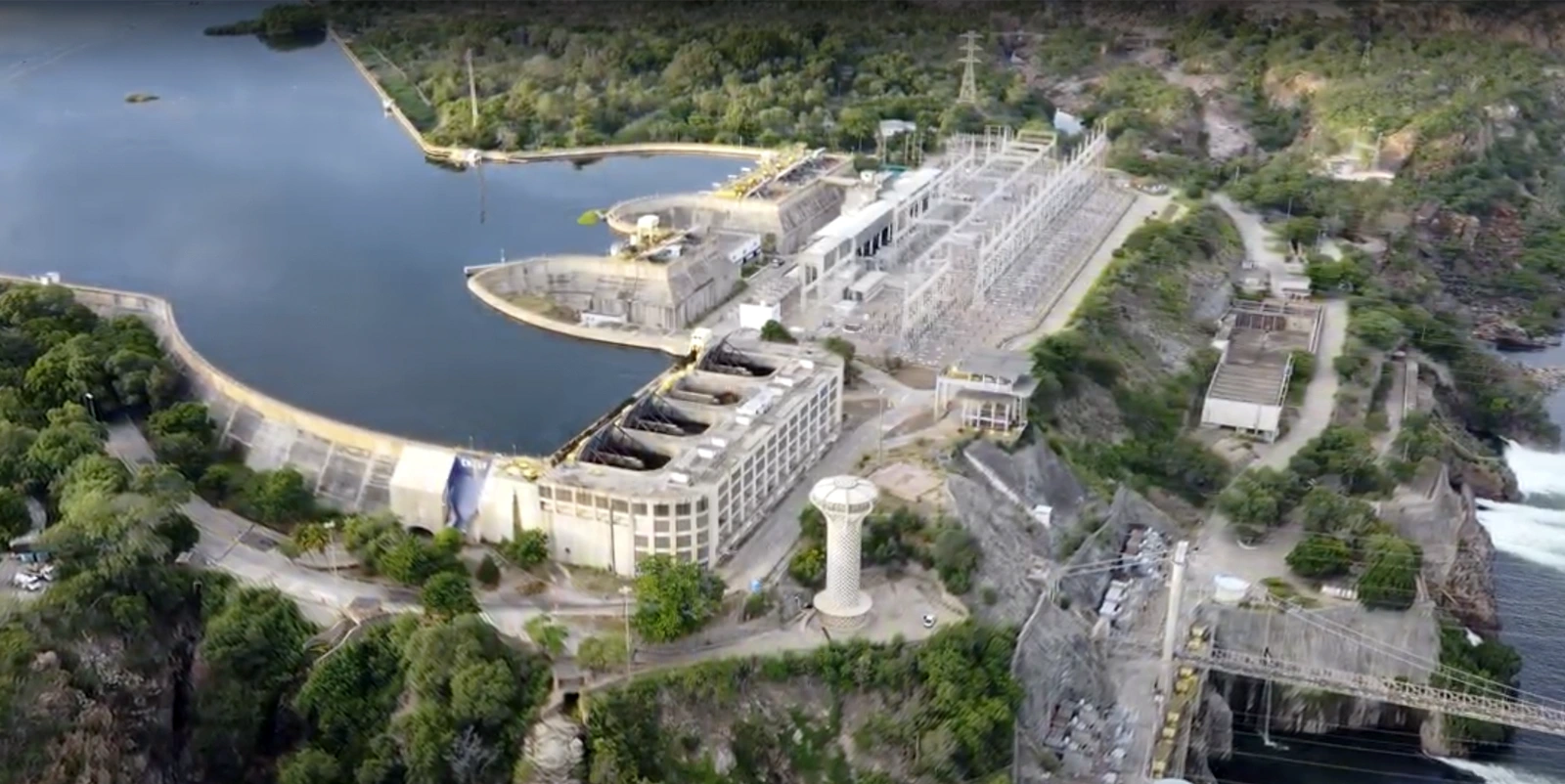 Vista aérea de um complexo hidrelétrico com barragem curva formando um reservatório, casa de força e subestação elétrica adjacente, cercadas por vegetação e relevo rochoso, com canais de escoamento de água visíveis nas laterais.
