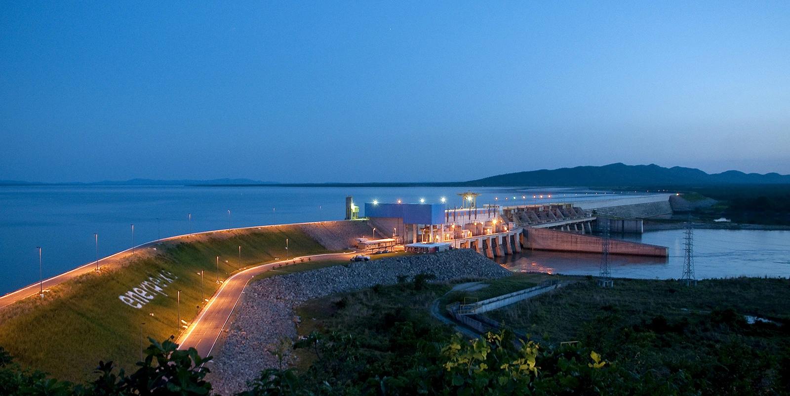 Usina hidrelétrica ao entardecer, com barragem iluminada por luzes, estrada curva à esquerda e grande corpo d’água ao fundo sob céu azul.