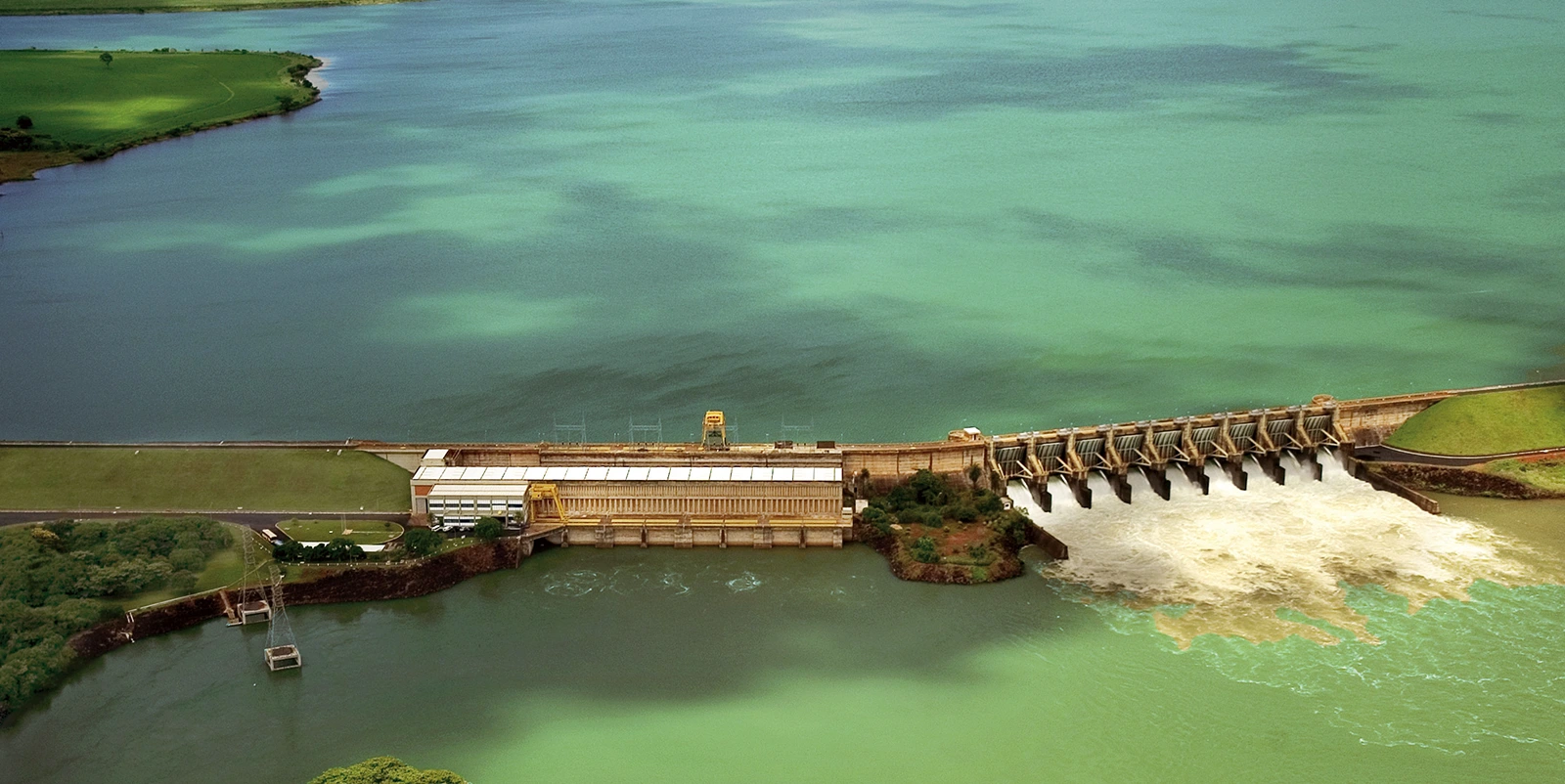 Vista aérea de uma usina hidrelétrica com barragem longa separando um grande reservatório de águas azul-esverdeadas e um canal a jusante com forte fluxo, incluindo casa de força retangular ao centro e vertedouro com múltiplas comportas abertas à direita, cercados por áreas verdes, margens gramadas e estruturas operacionais ao redor.