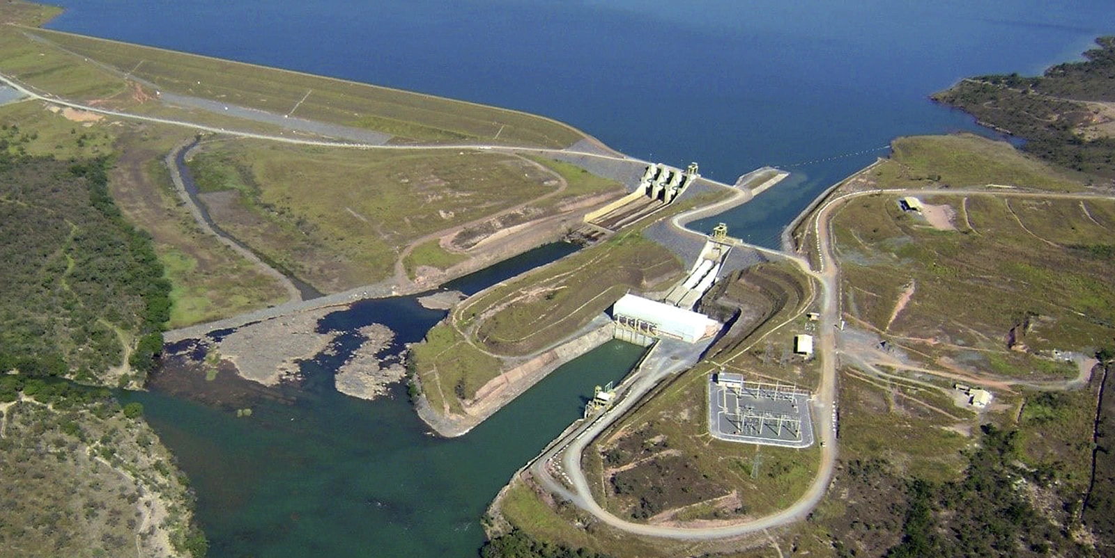Vista aérea de um complexo hidrelétrico com barragem, canal de adução e casa de força conectando diferentes níveis de água, cercado por relevo suave, estradas de acesso e reservatório amplo ao fundo.