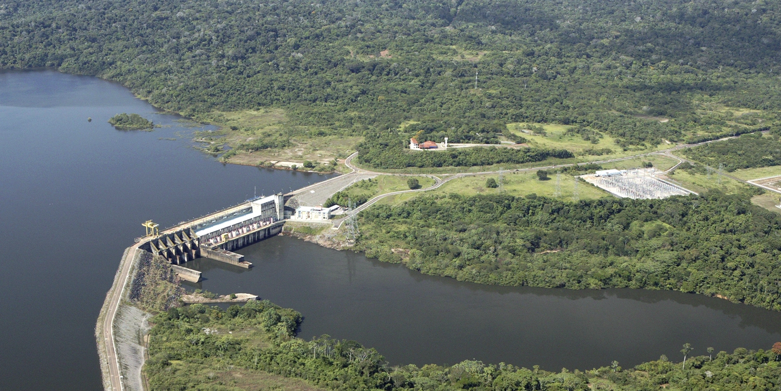 Vista aérea de uma usina hidrelétrica de pequeno porte com barragem e casa de força compacta junto ao canal, conectada a uma subestação elétrica próxima, cercada por vegetação densa e reservatório amplo que se estende ao fundo em meio a área florestal.