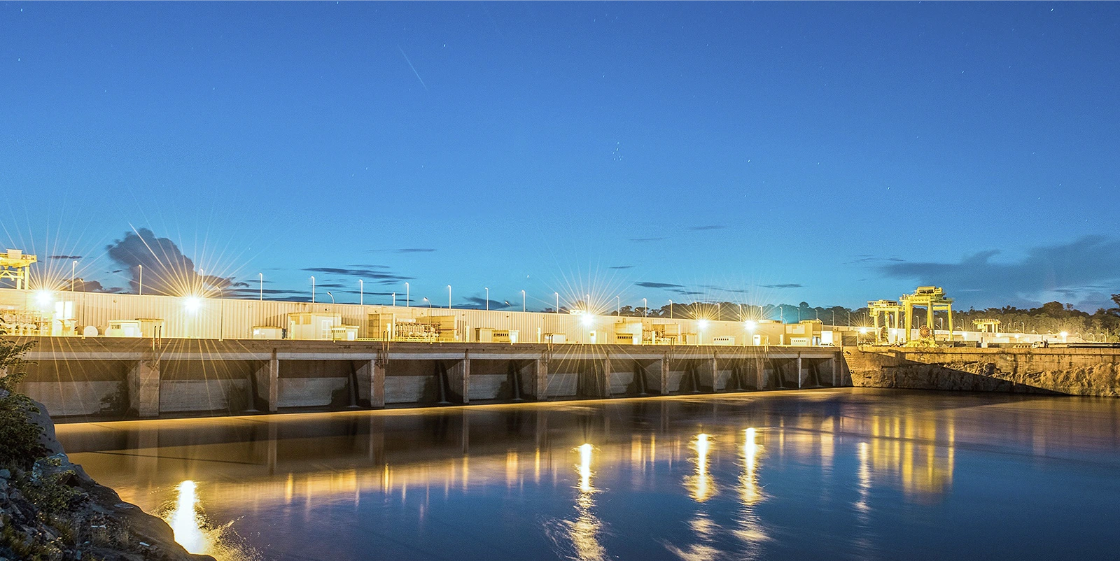 Vista noturna de uma usina hidrelétrica iluminada, com barragem e casa de força refletindo luzes douradas na água calma do rio, sob céu azul profundo ao entardecer.