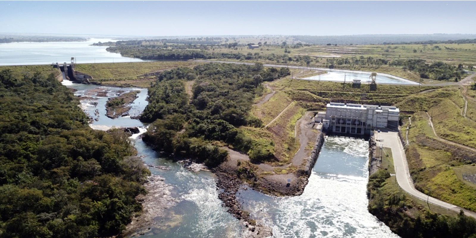Vista aérea de um sistema hidrelétrico integrado com barragem ao fundo, reservatório extenso e casa de força posicionada à direita liberando água para um canal a jusante, cercado por áreas de vegetação densa, relevo suave, estradas de acesso e estruturas operacionais distribuídas ao longo do complexo, sob céu claro.