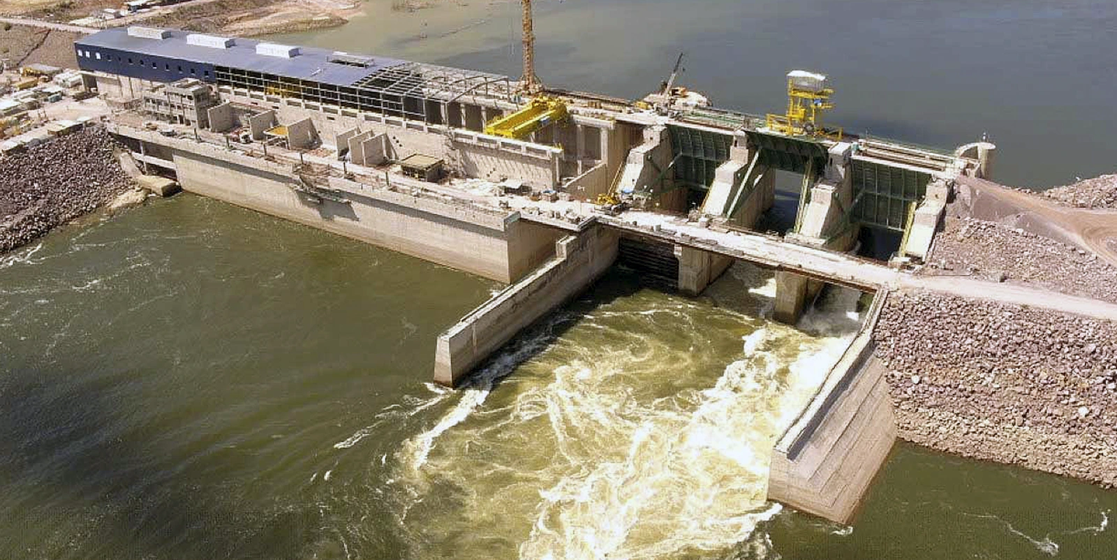 Usina hidrelétrica em operação vista de cima, com comportas liberando água, estruturas industriais e guindaste ao lado de um grande reservatório.