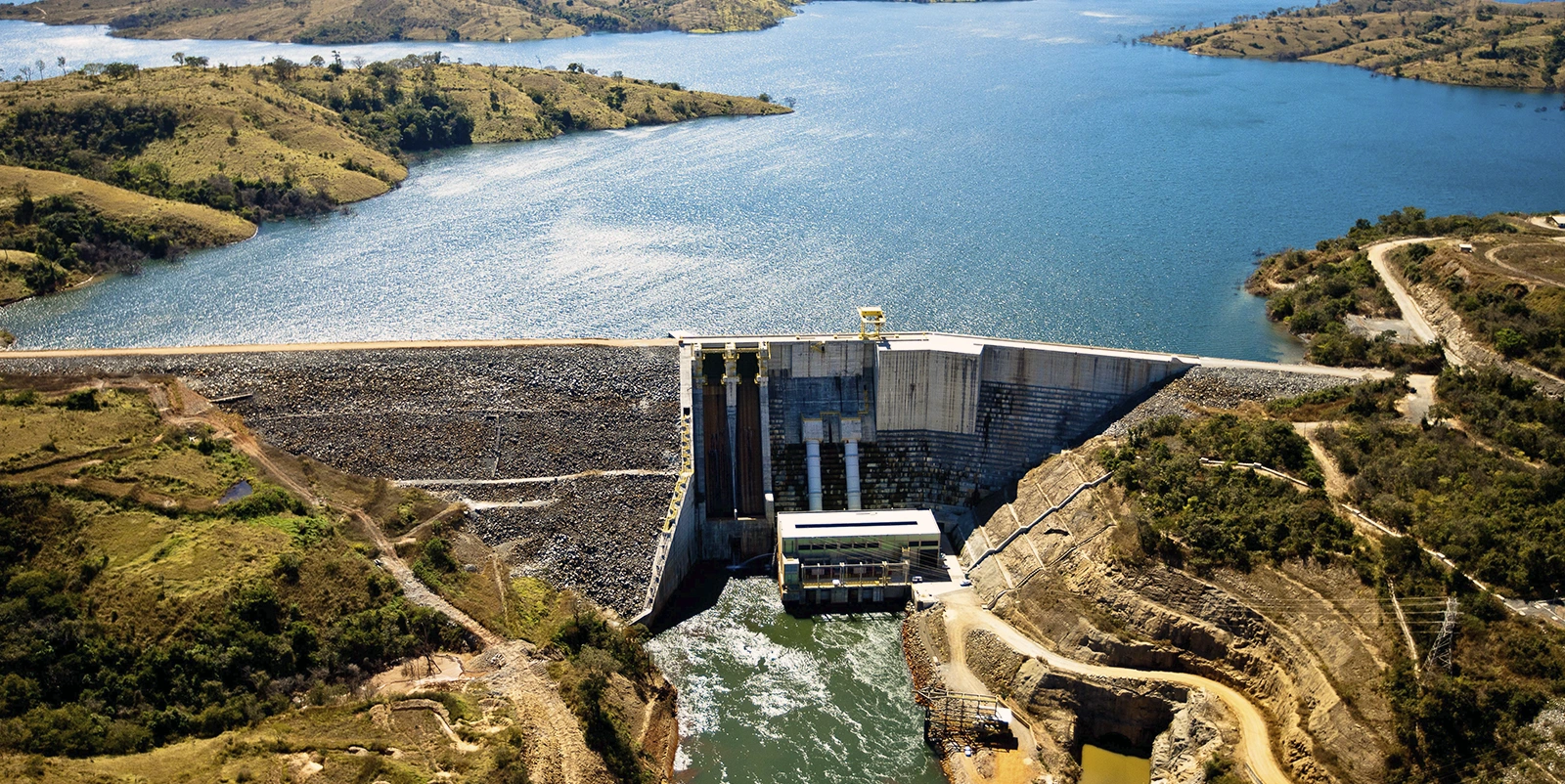 Vista aérea de uma usina hidrelétrica com barragem de enrocamento e estrutura central de concreto, conectada a uma casa de força na base, onde a água escoa para o canal a jusante, cercada por reservatório amplo, colinas com vegetação esparsa e estradas sinuosas ao redor.