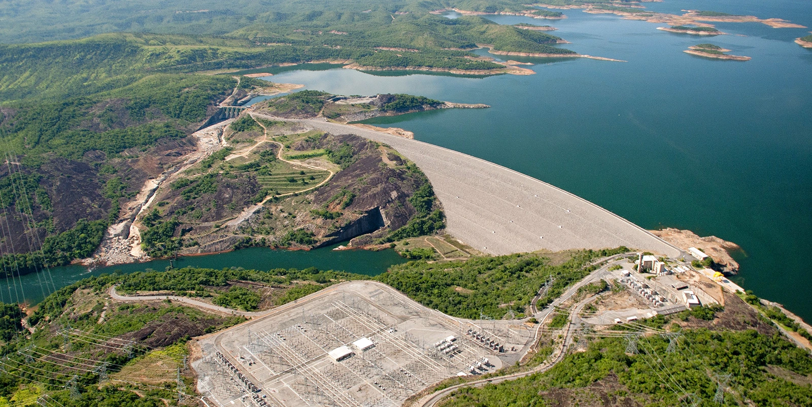 Vista aérea de uma barragem hidrelétrica extensa formando um grande reservatório com ilhas e margens irregulares, cercada por áreas de vegetação, relevo montanhoso e estrada de acesso ao longo da estrutura.