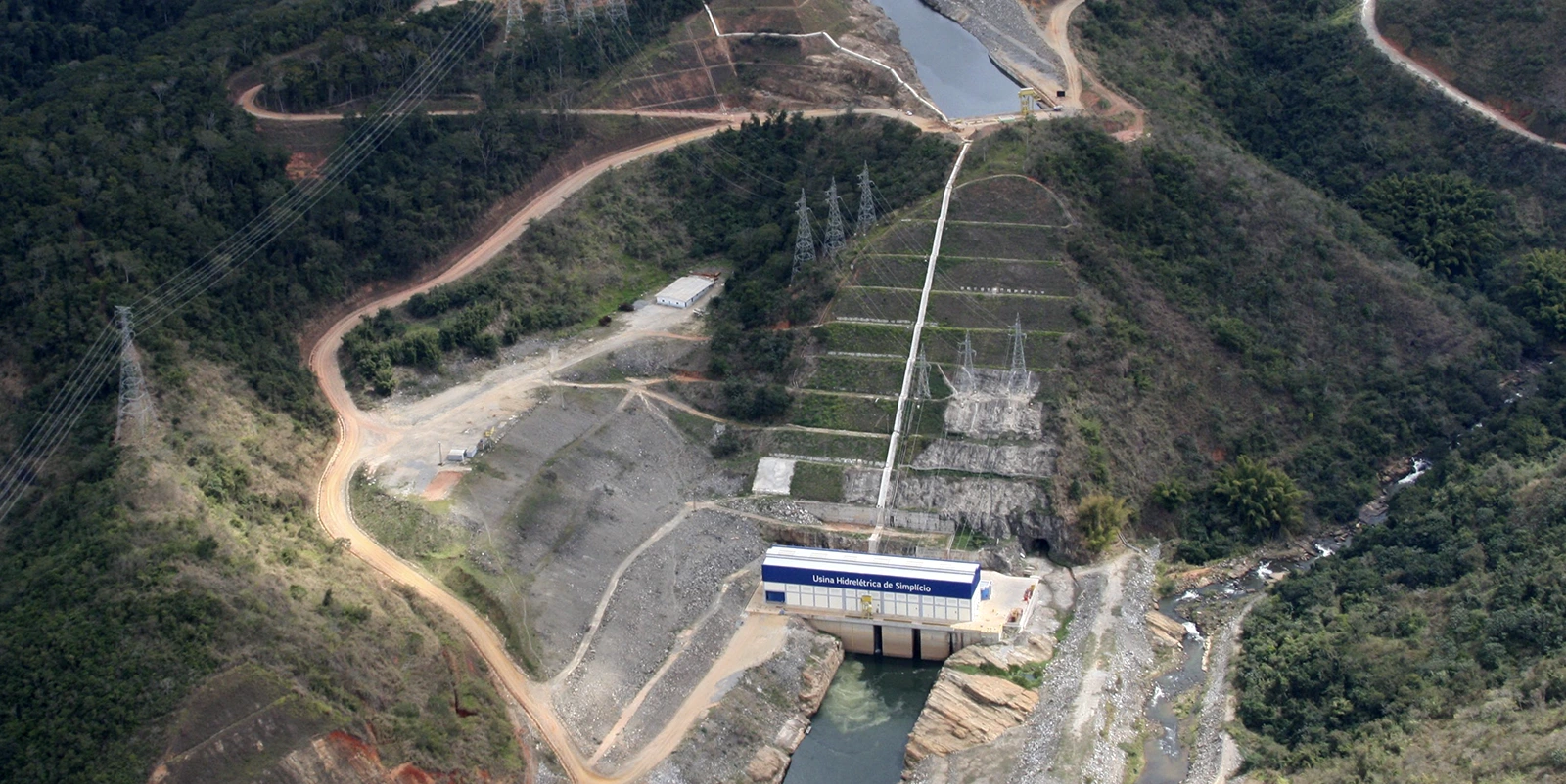 Vista aérea de um pequeno aproveitamento hidrelétrico em vale cercado por vegetação densa, com casa de força identificada como “Usina Hidrelétrica de Simplício” posicionada sobre um canal estreito, interligada por estradas de terra, linhas de transmissão e estruturas elétricas distribuídas ao longo das encostas.