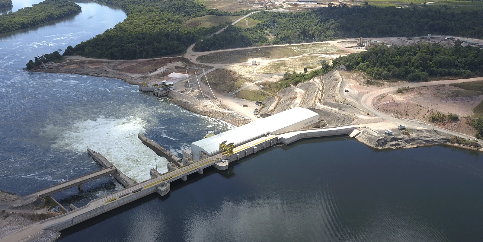 Vista aérea de um complexo hidrelétrico compacto com barragem baixa e casa de força alongada, onde a água escoa em forte correnteza para um canal lateral, cercado por áreas em terraplenagem, estradas de acesso, vegetação densa nas encostas e um rio mais amplo ao fundo.