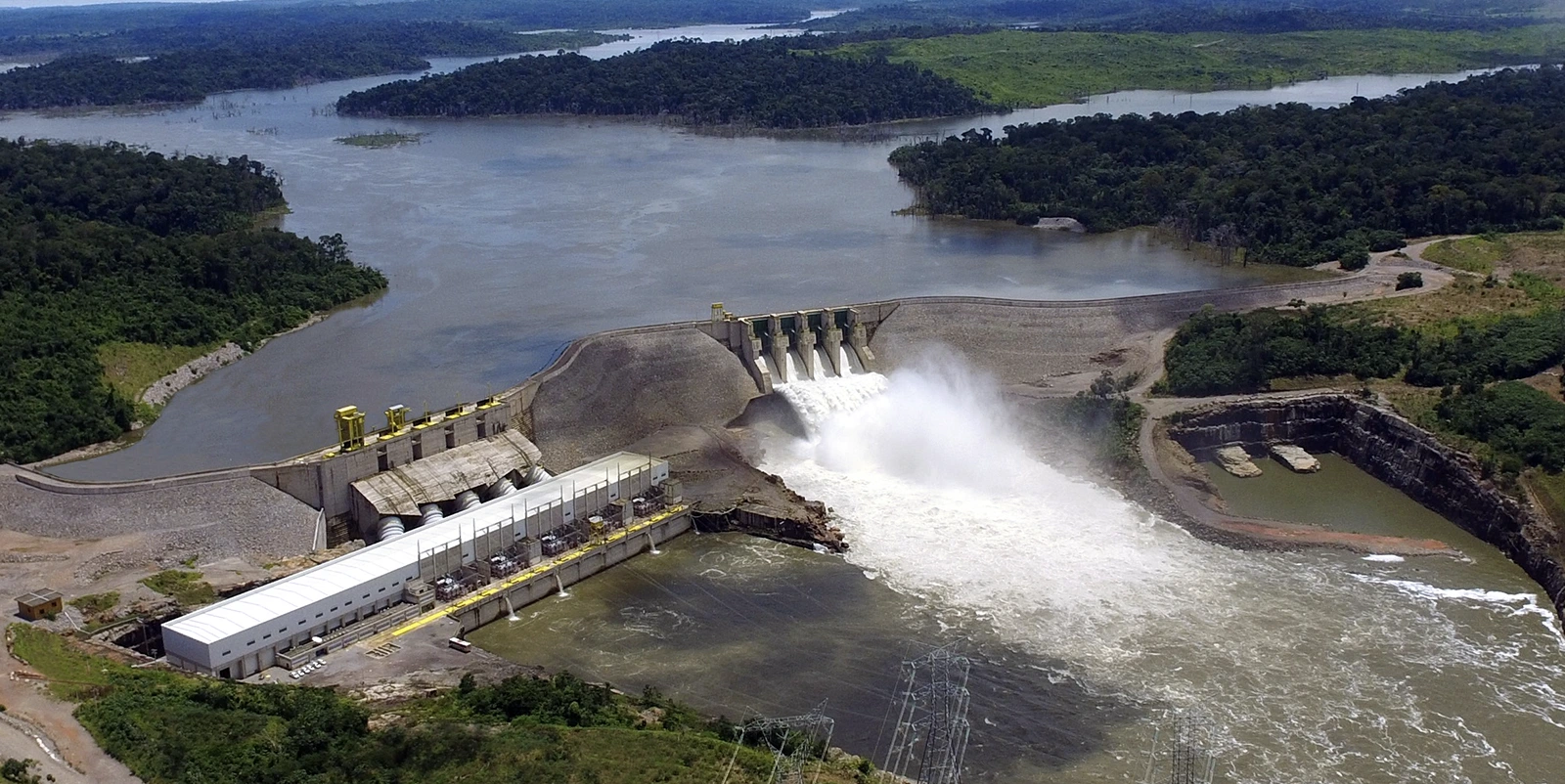 Vista aérea de uma usina hidrelétrica com barragem de enrocamento e casa de força alongada, onde o vertedouro libera grande volume de água, formando espuma no canal de jusante, cercada por vegetação densa e reservatório que se estende ao fundo.
