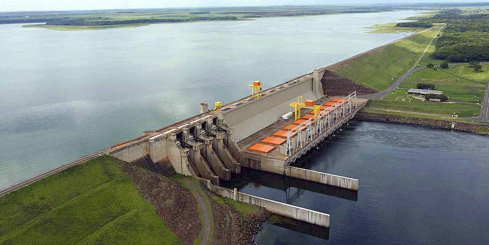 Barragem hidrelétrica vista de cima durante o dia, com vertedouros, estruturas industriais e grande reservatório de água ao redor, cercado por áreas verdes.