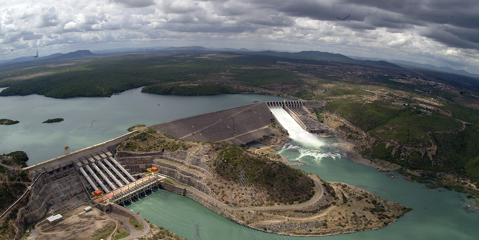 Vista aérea ampla de um complexo hidrelétrico com duas estruturas de barragem — uma com vertedouro liberando grande volume de água e outra com vários condutos inclinados — conectadas por relevo montanhoso, formando reservatórios em níveis diferentes, cercadas por vegetação esparsa e paisagem extensa sob céu carregado de nuvens.
