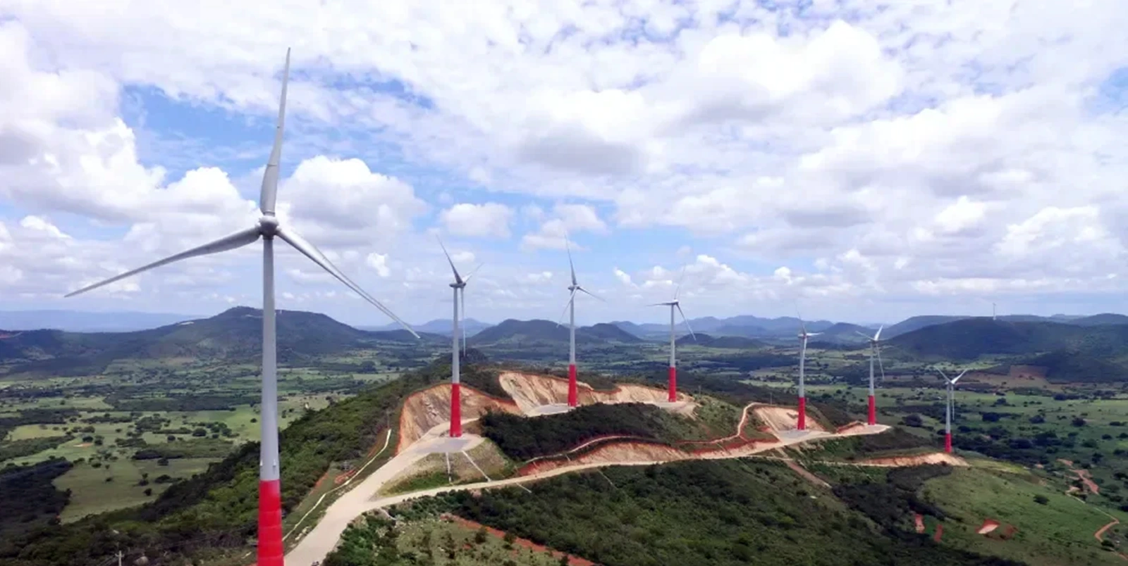 Parque eólico com várias turbinas alinhadas sobre morros verdes, sob céu parcialmente nublado.