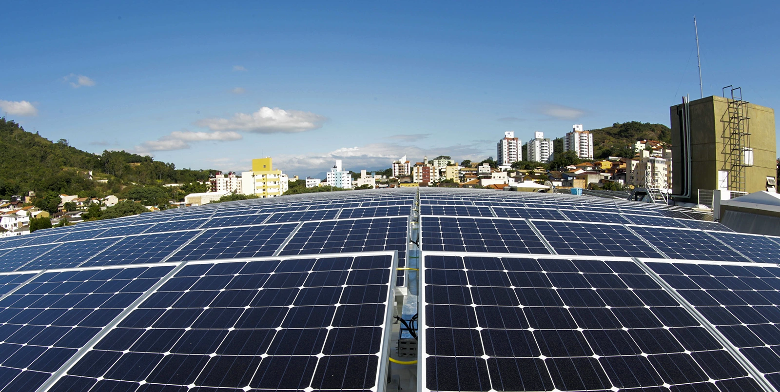 Painéis solares instalados em telhado urbano com prédios residenciais ao fundo sob céu claro.