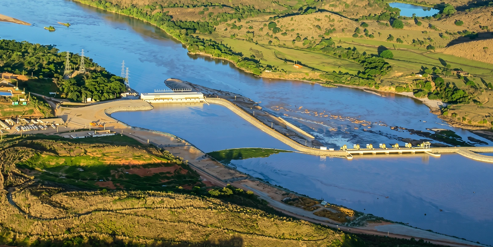 Vista aérea de uma usina hidrelétrica com barragem sobre um rio largo, cercada por colinas verdes e áreas rurais, sob luz natural.