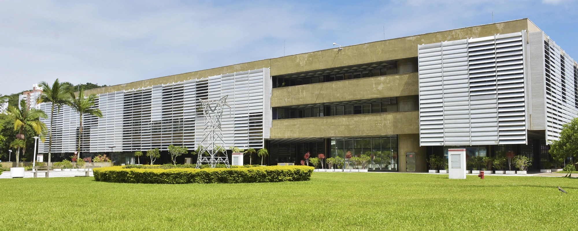Edifício moderno com fachada de brises metálicos e concreto, cercado por gramado e árvores, com torre de energia elétrica decorativa em frente.