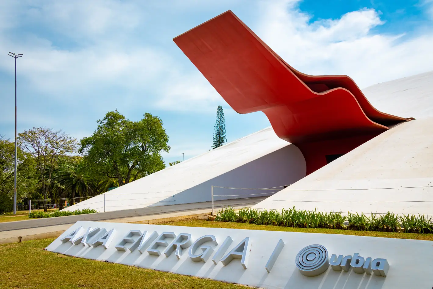 Fachada do Auditório Ibirapuera, em São Paulo, com arquitetura moderna de linhas brancas e destaque para a marquise vermelha ondulada característica do projeto de Oscar Niemeyer, cercada por áreas verdes.