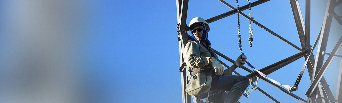 Trabalhador usando capacete e óculos de proteção está suspenso por cordas e correntes em uma estrutura metálica alta, realizando trabalho em altura contra um céu azul limpo.