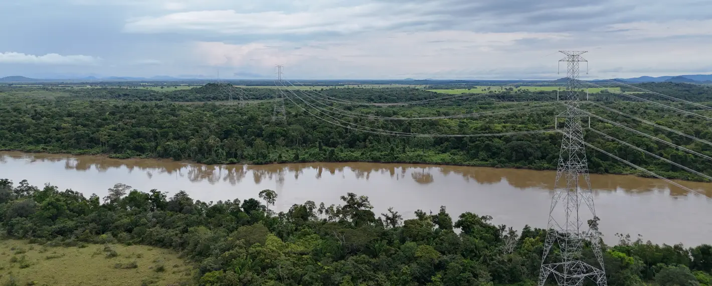 Vista aérea do linhão Manaus-Boa Vista atravessando área de floresta e rio na Amazônia. Torres de transmissão de energia se destacam na paisagem, reforçando a integração elétrica e a segurança energética em Roraima.