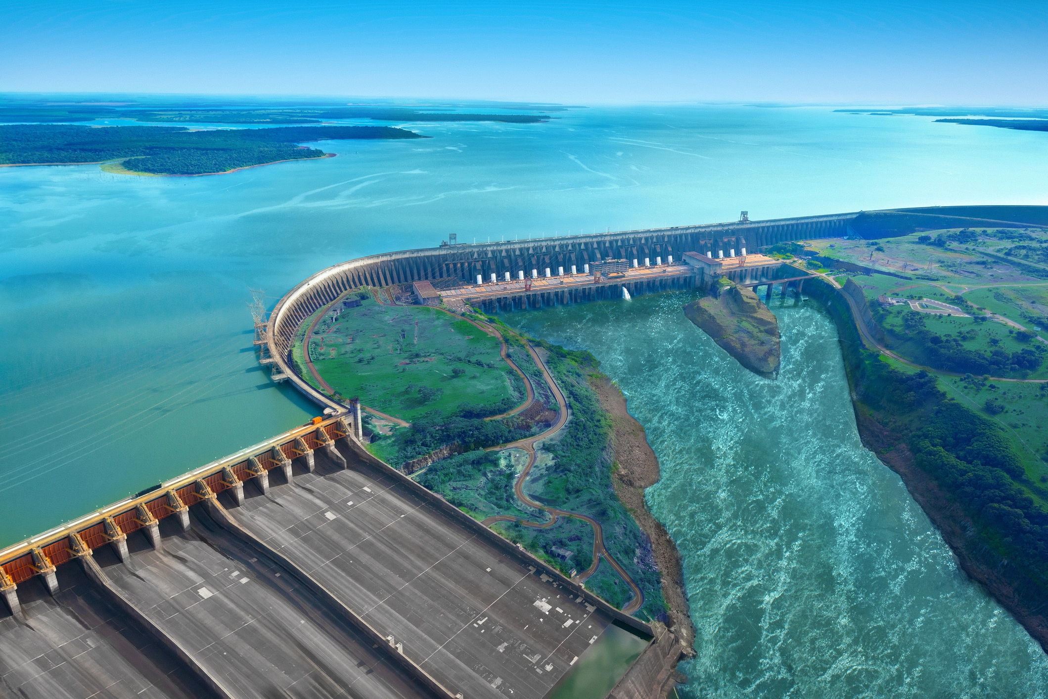 Vista aérea de uma grande usina hidrelétrica com barragem, reservatório e vertedouro, cercada por água e áreas verdes.
