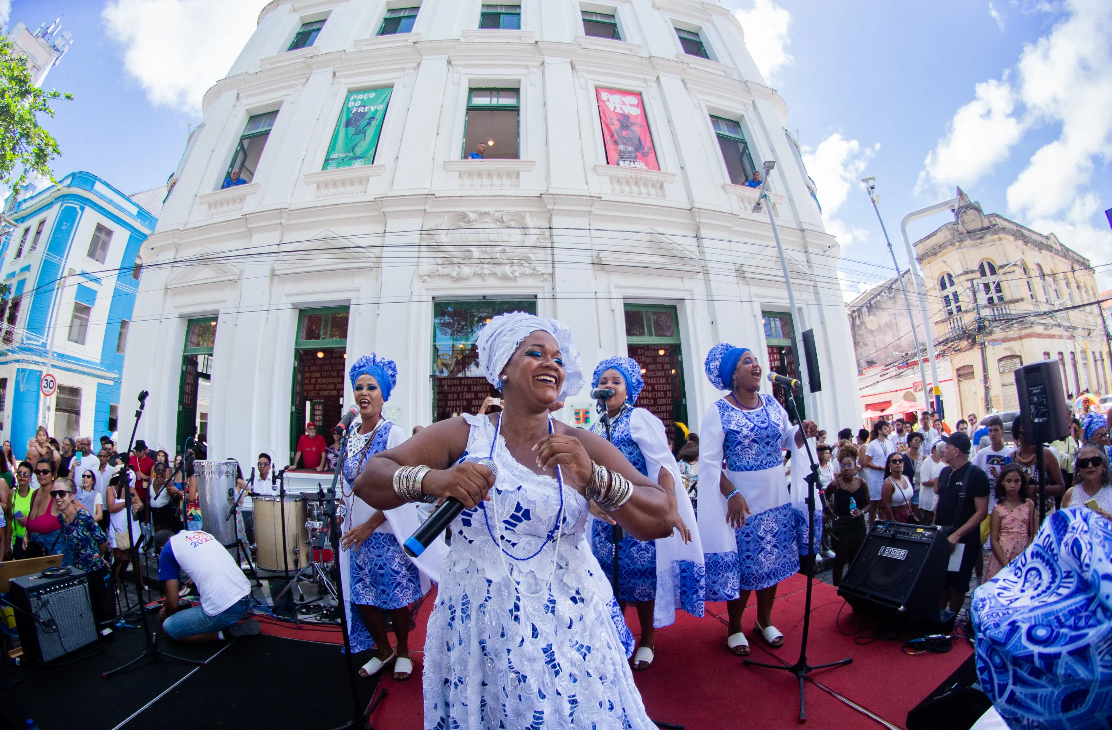Grupo de mulheres em trajes tradicionais brancos e azuis se apresenta cantando e dançando em uma rua movimentada, diante de um prédio histórico, durante uma celebração cultural.